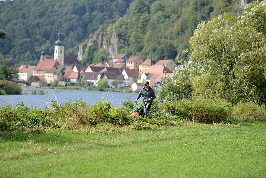 Ein Mann mäht einen Uferrandstreifen an einem See in dessen Hintergrund ein Dorf vor einem steilen bewaldeten Hang zu sehen ist.
