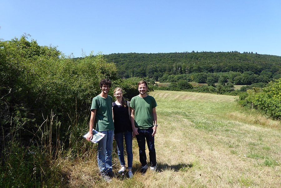 Drei Personen stehen vor einer Hecke in der Landschaft.