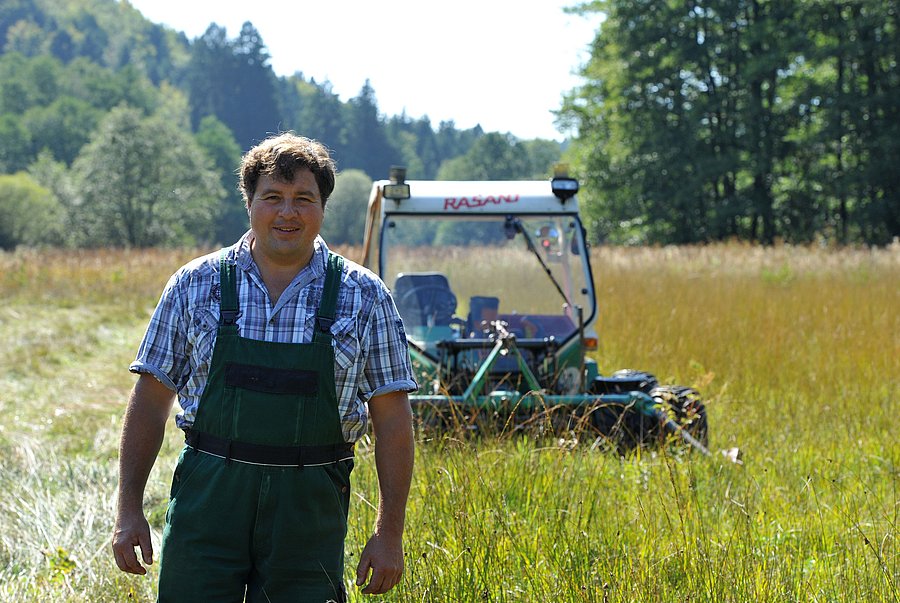 Landwirt Hans Maurer steht in einer Streuwiese. Hinter ihm ist sein Mähtrac mit Zwillingsbereifung zu sehen.