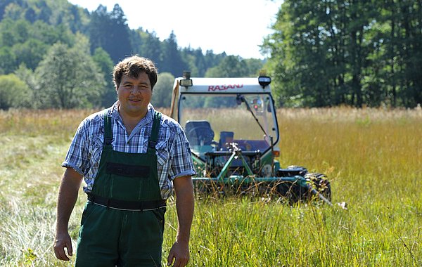 Landwirt Hans Maurer steht in einer Streuwiese. Hinter ihm ist sein Mähtrac mit Zwillingsbereifung zu sehen.