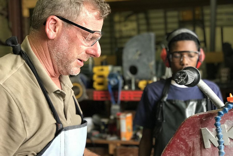 Ein Mann mit Schutzbrille steht in der Werkstatt an einer Maschine zur Holzbearbeitung. Ein Junge schaut ihm zu