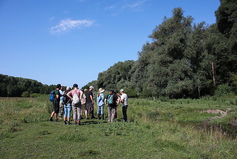 ©A. Schuller/DVL Gruppe von Menschen auf einer Wiese neben einem Bachlauf und hohen Bäumen