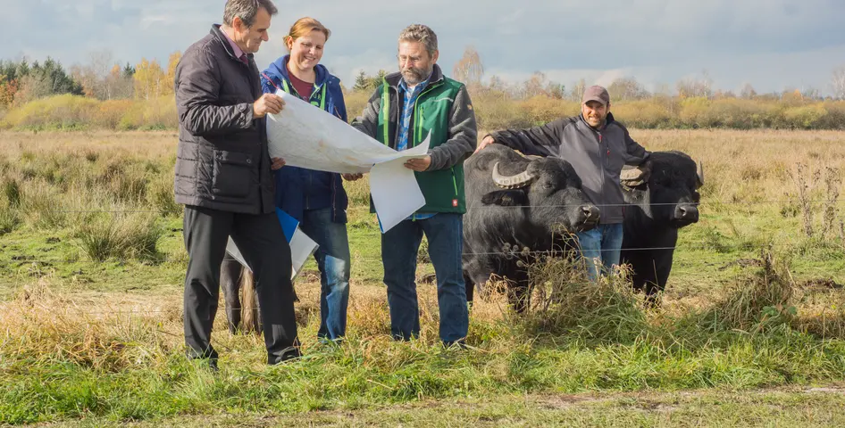 Drittelparitat im Schwäbischen Donaumoos ©Erich Herrmann Bürgermeister, Landwirtin und der Geschäftsführer der ARGE Schwäbisches Donaumoos stehen auf einer Weide im Moor. Sie halten eine Landkarte. Im Hintergrund ist ein Landwirt mit seinen Wasserbüffeln und zu sehen.