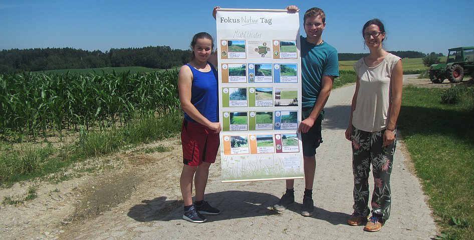 Drei Personen stehen auf einem Feldweg und halten ein Plakat hoch.