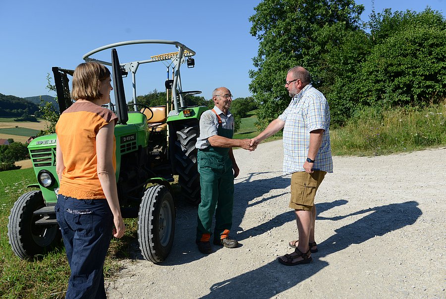 Ein Landwirt und ein Berater stehen neben einem Traktor und schütteln sich die Hand. Eine Beraterin schaut dabeizu.